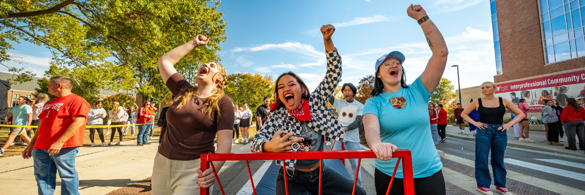 Students cheer and celebrate during Ball State University's Homecoming Bed Race, standing behind a red bed frame on Riverside Avenue while spectators line the street on a sunny fall day.