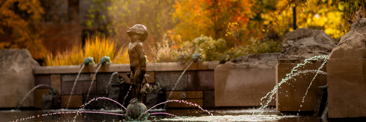 The Frog Baby bronze statue standing atop a sphere inside a fountain, with water spraying around its base. Small frog spouts stream water in the background set against vibrant yellow and orange fall trees illuminated by soft sunlight.
